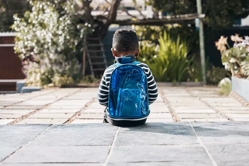 Image: a sad young child with a backpack sitting, seen from behind
