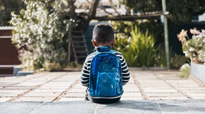 a sad young child with a backpack sitting, seen from behind