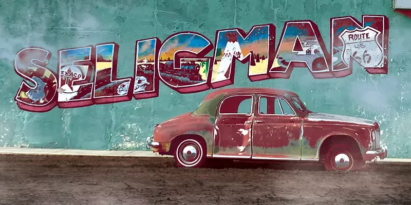 Image: a rusted car in front of a wall with a postcard-style "Seligman" on the side
