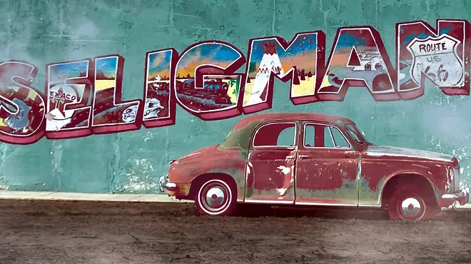 a rusted car in front of a wall with a postcard-style "Seligman" on the side