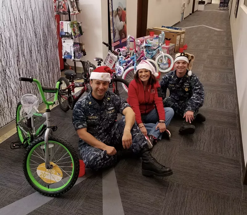 Image: Margy Bons, center, sits with two service members as they prepare for a bicycle raffle during Military Assistance Mission’s Operation Holiday Open House.