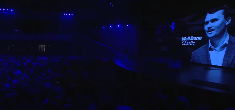 Image: a darkened auditorium with a screen featuring charlie kirk and the words "well done charlie"