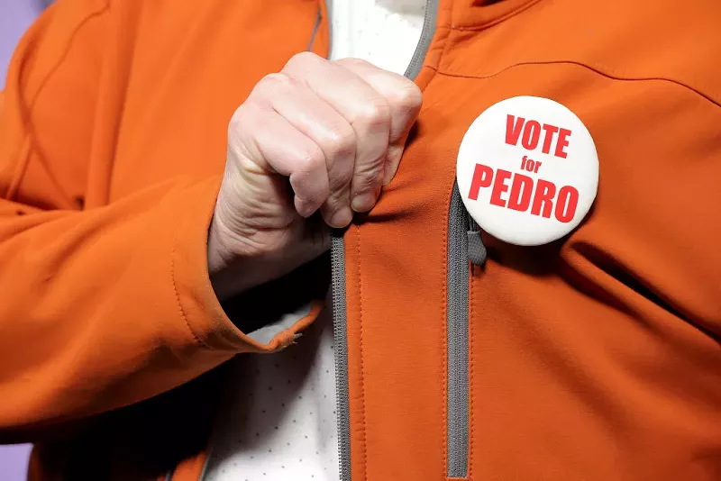 Image: a hand grabs the lapel of an orange jacket that features a "vote for pedro" pin