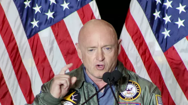Mark Kelly speaks in a military jacket in front of the U.S. flag.