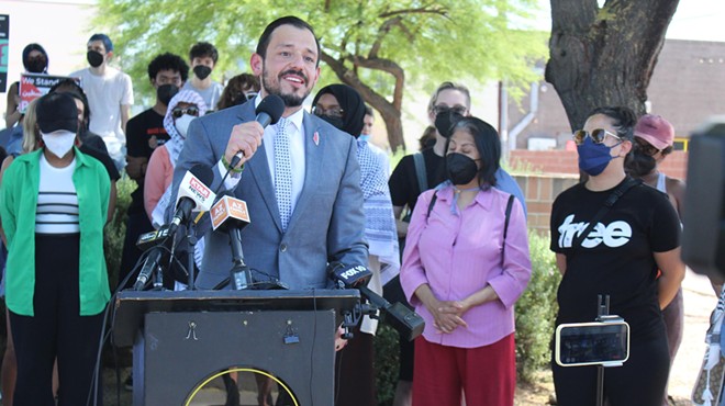 A man in a suit stands before a podium with a sign for "Mass Liberation AZ" and speaks into TV microphones. People in face masks stand behind him.