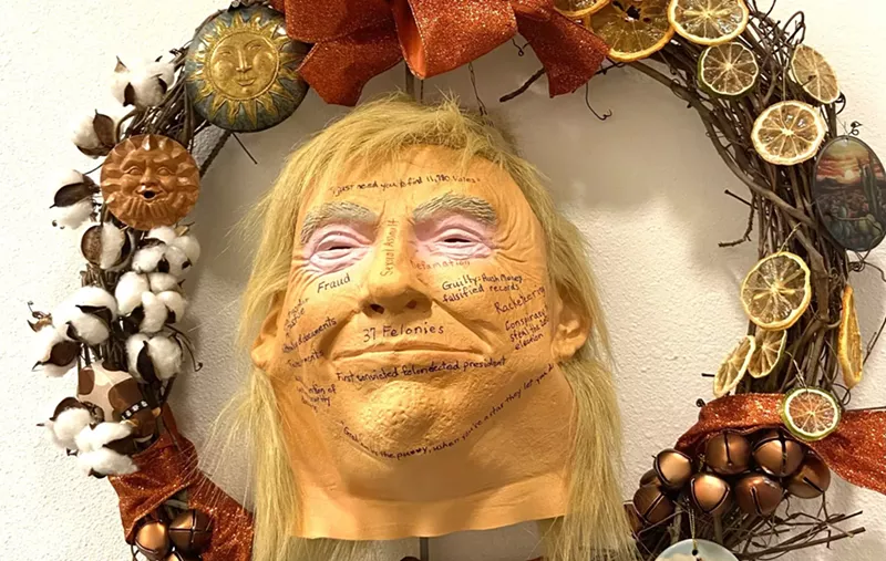 Image: a wreath featuring a rubber trump mask with phrases like "defamation" and "sexual assault" written on its face
