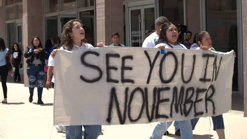 Image: Three people march holding a banner that says, "See you in November"