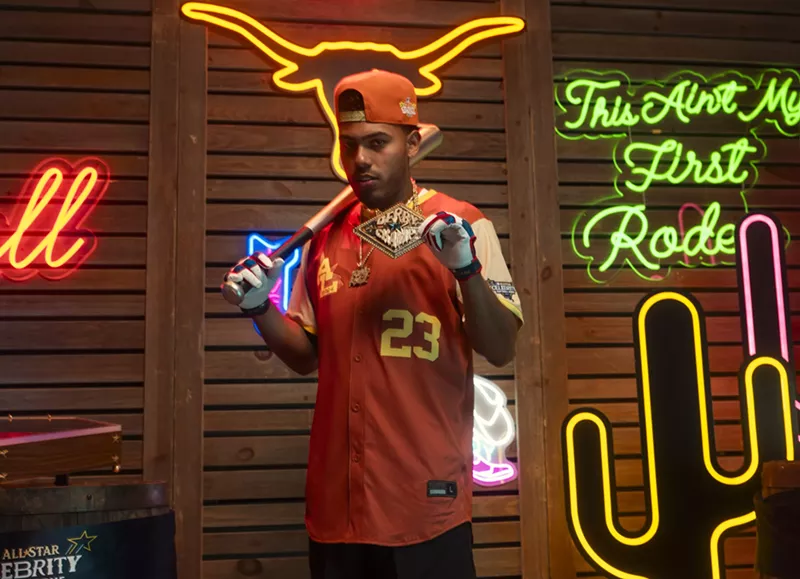 Image: A man in a baseball jersey in front of a wall of neon signs.