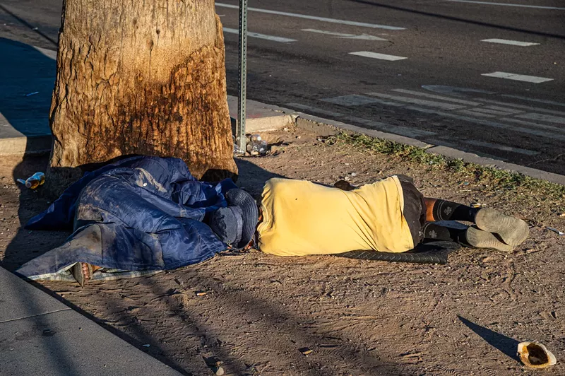 Image: a unhoused man in a yellow shirt laying, back to the camera, at the base of a tree