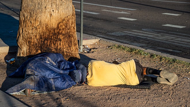 a unhoused man in a yellow shirt laying, back to the camera, at the base of a tree