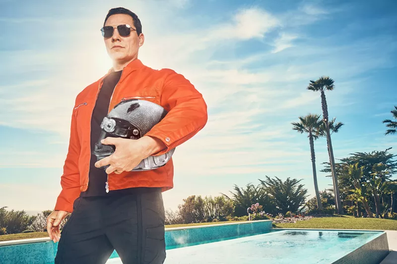 Image: A man in an orange jacket holding a motorcycle helmet.