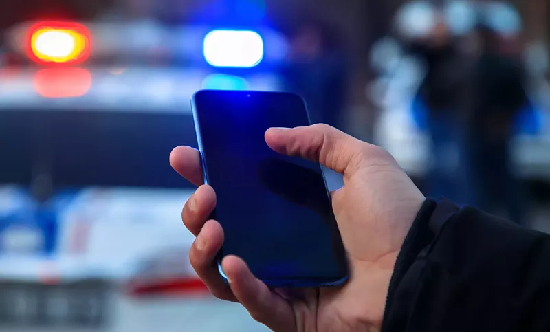 Image: a hand holds a phone in front of a cop car with flashing lights