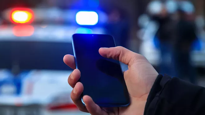 a hand holds a phone in front of a cop car with flashing lights