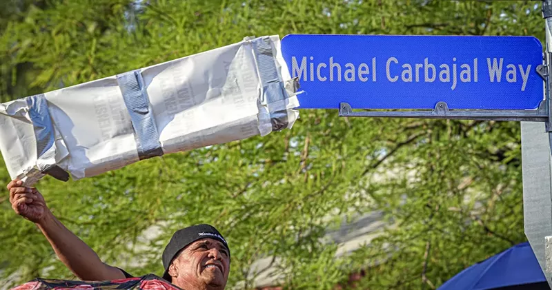 Image: A man pulling a cover of a street sign that reads "Michael Carbajal Way"