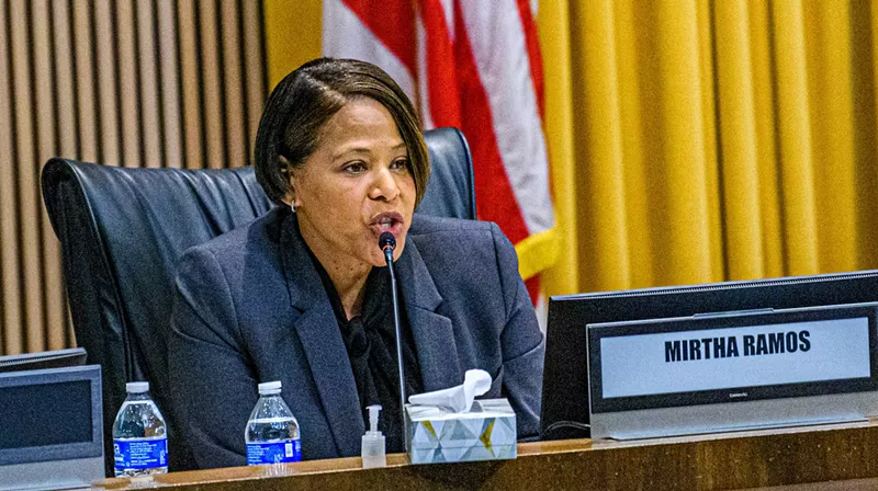 Image: a woman speaks into a microphone in a meeting room setting