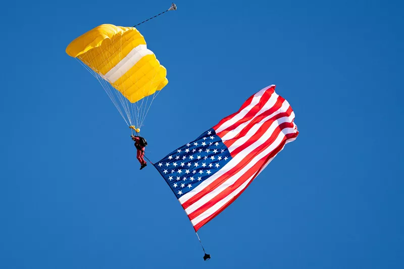 Image: a skydiver with a yellow parachute and the American flag trailing below him