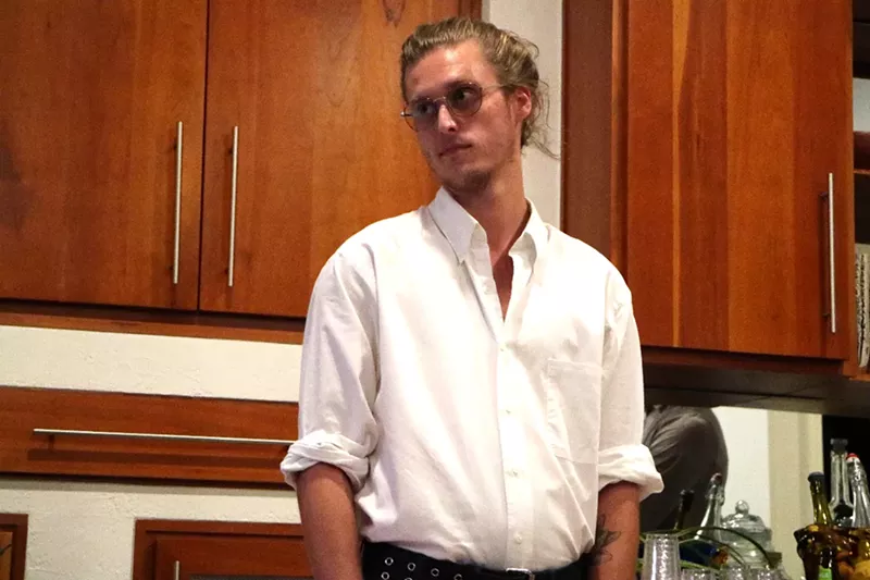 Image: a young man in glasses with his blond hair up in a bun
