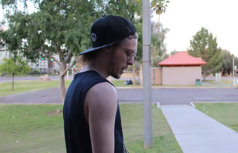 Image: A man in glasses, a backward black hat and a black tank top looks at the ground