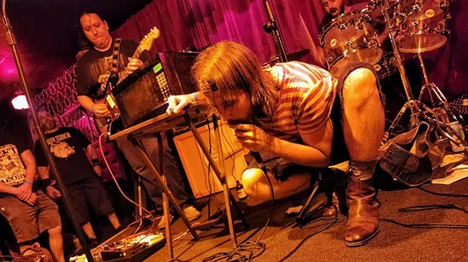 A long-haired man crouches on a stage while holding and singing into a microphone.
