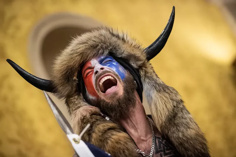 Image: the qanon shaman, with a buffalo headdress and red-white-and-blue facepaint