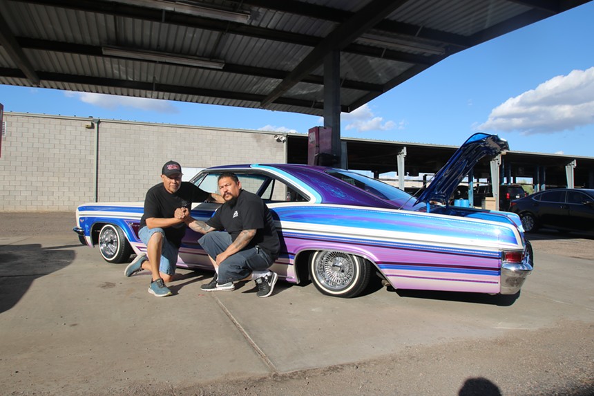 Two men crouch in front of a colorful old car with the trunk open.