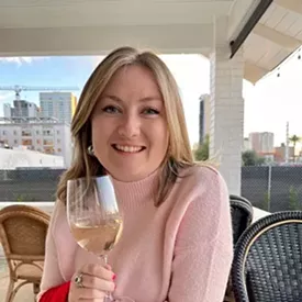 A woman with blonde hair smiles and holds a glass of white wine.
