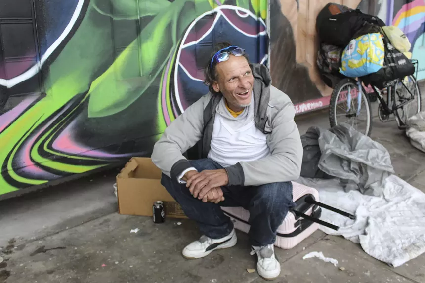 An unsheltered man sits in front of a colorful mural.