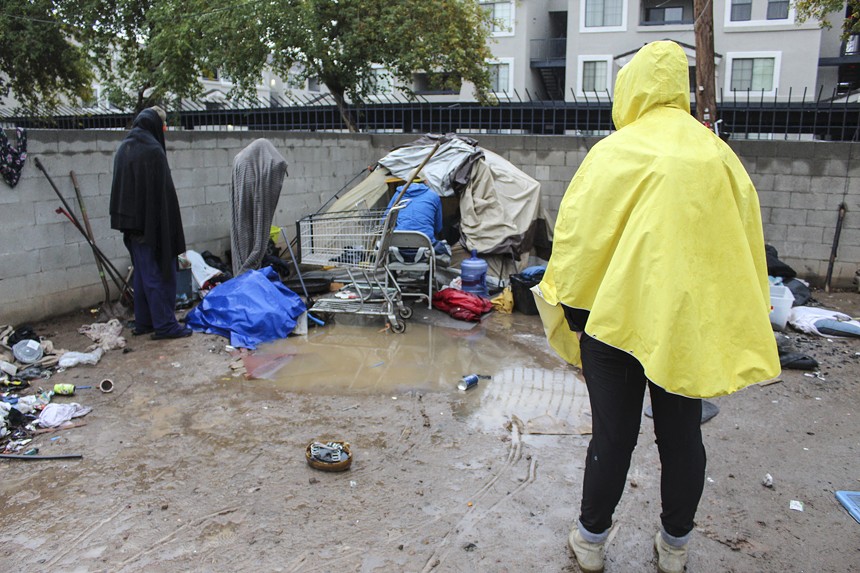 A man in a rain poncho watches as another man speak to people living in tents.