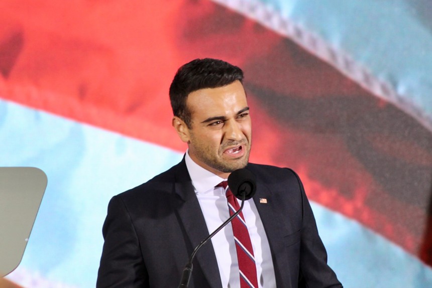 A man in a black suit and red striped tie standing at a microphone in front of a huge American flag backdrop.