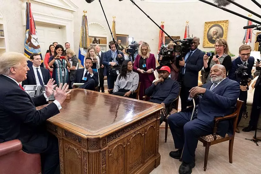 Kanye West sits, in a red MAGA hat and surrounded by others, in front of the desk of Donald Trump in the Oval Office.
