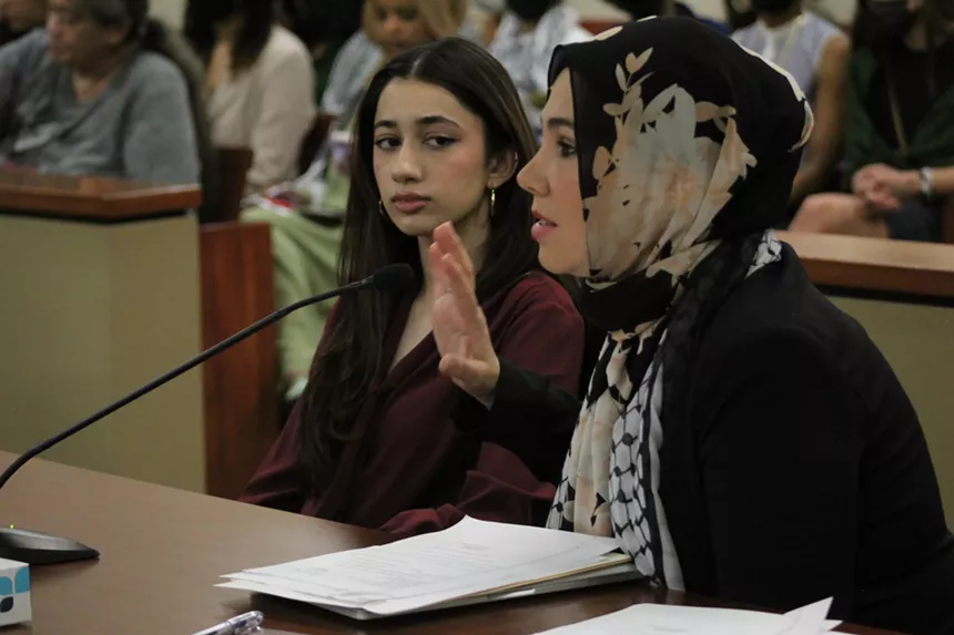 A woman in a headscarf raises her hand while speaking into a courtroom microphone as another woman looks on.