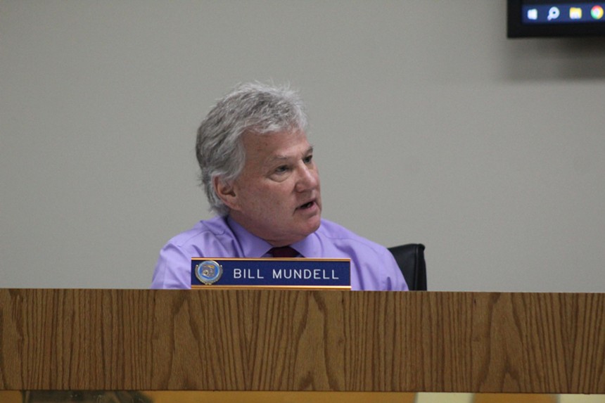 A man in front of a nameplate that reads "Bill Mundell" speaks into a microphone.