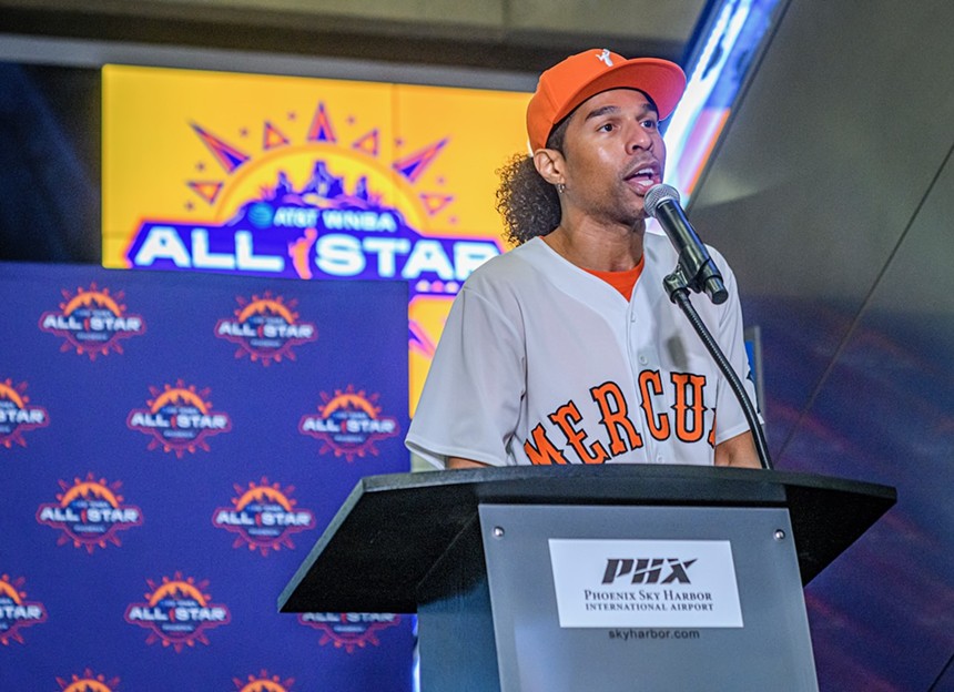 A man in an orange hat and a white, baseball-style "Mercury" jersey speaks at a podium