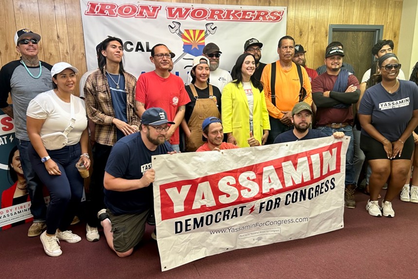 A woman in a yellow blazer poses with volunteers, all holding a sign that says "YASSAMIN"