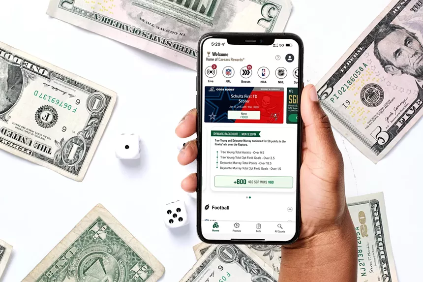 A hand holds a phone showing the Caesars Sportsbook app. Cash floats around in the background.
