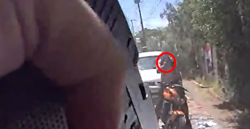 In the foreground is part of a hand holding a handgun. Beyond is a blurry image of a man on a motorcycle pointing something at the viewer. A red circle has been drawn over the man's hand.