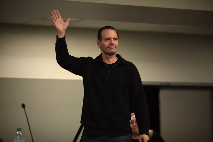 A middle-aged man in black clothing waves to a crowd.