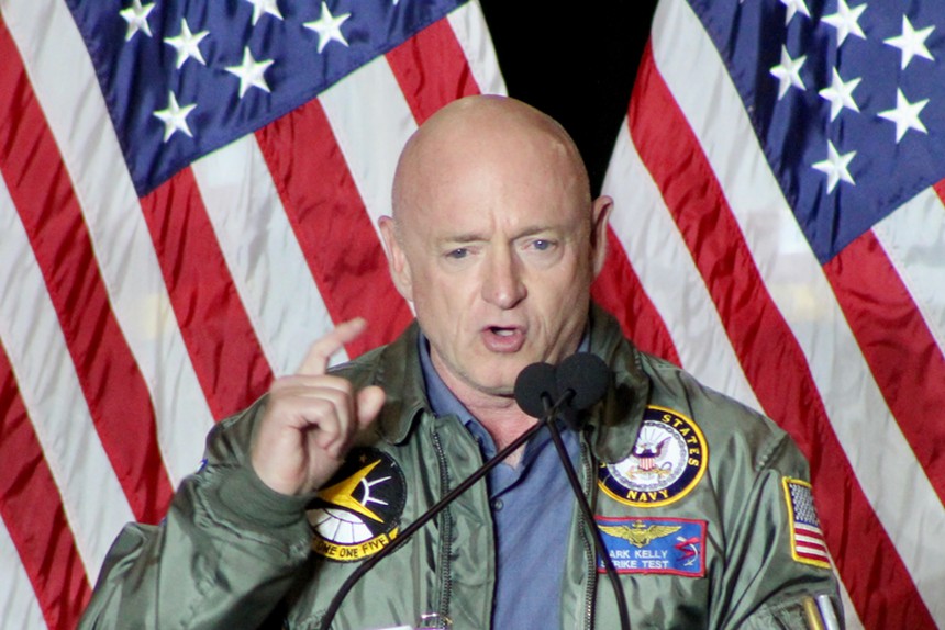 Mark Kelly speaks in a military jacket in front of the U.S. flag.