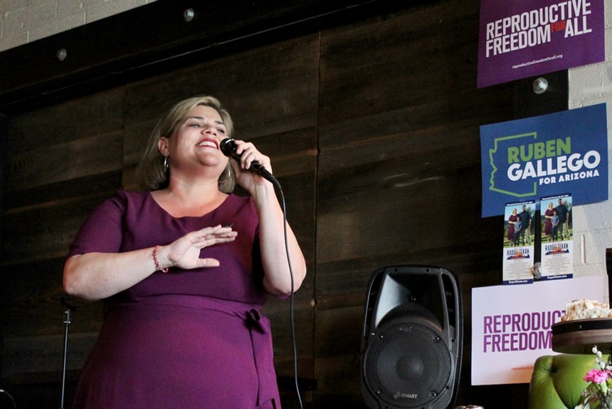 A Latina woman in a purple/maroon dress speaks into a microphone