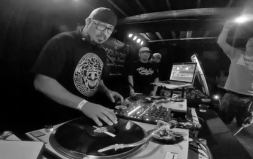 A Latino man DJs on a pair of turntables in a nightclub.