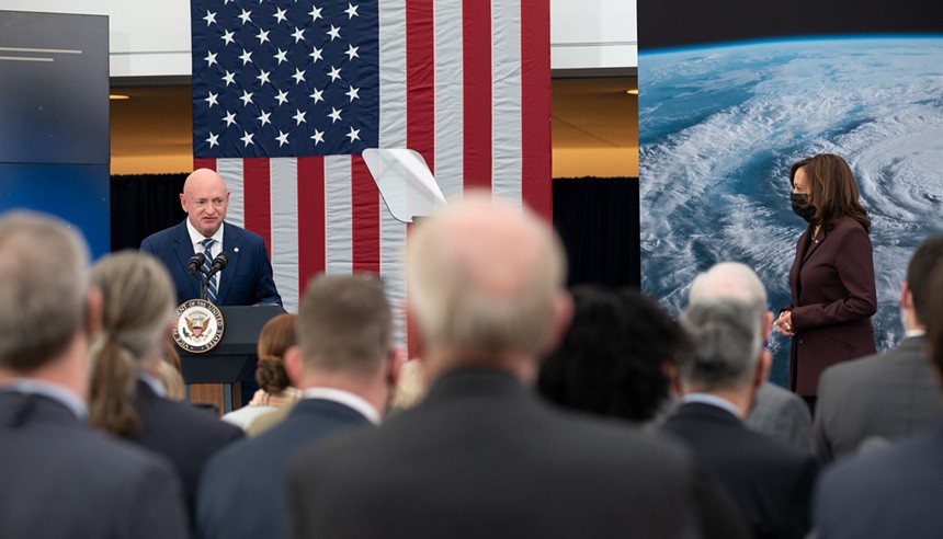 Mark Kelly at a podium in front of an American flag. To the side, Kamala Harris stands wearing a face mask