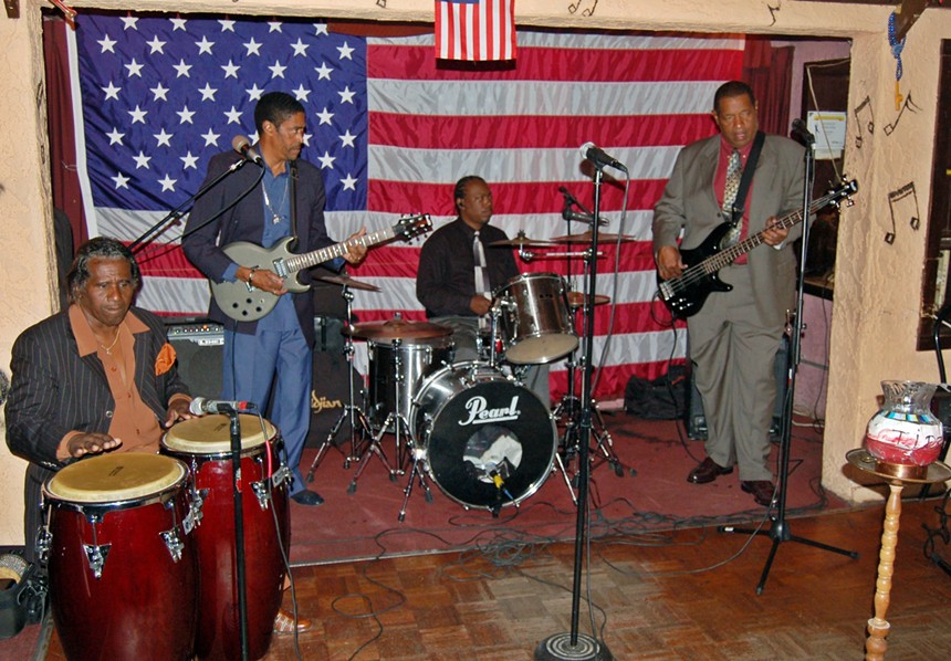 An R&B band performs in front of a large American flag inside a bar.