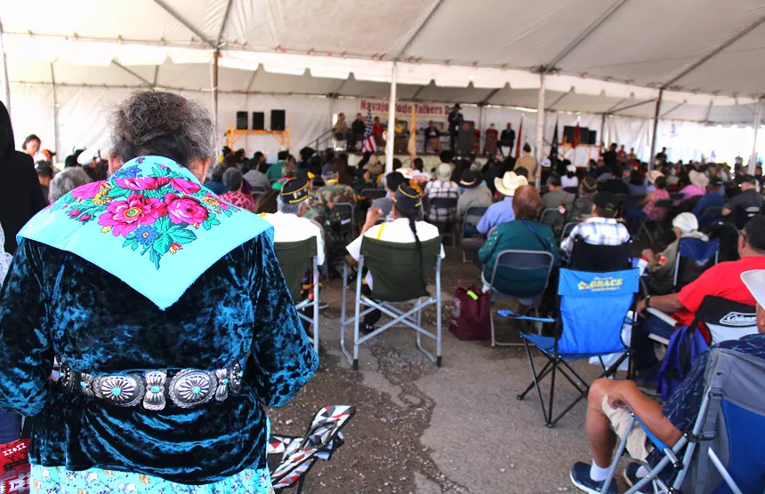 People sit under a tent watching a person speak at a podium.