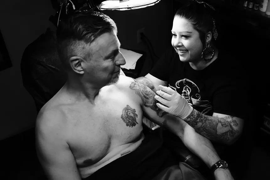 A black and white photo of a woman tattooing a man.