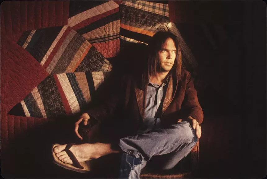 A man with long hair sits in front of a quilt.
