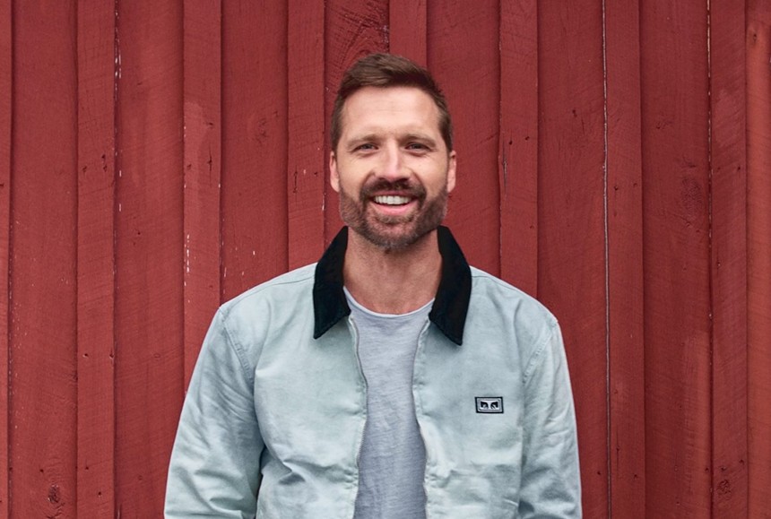 A bearded man in a gray jacket stands in front of a wall.