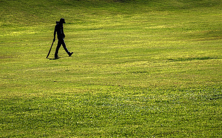 a man in silhouette drags a cricket bat on a grassy field