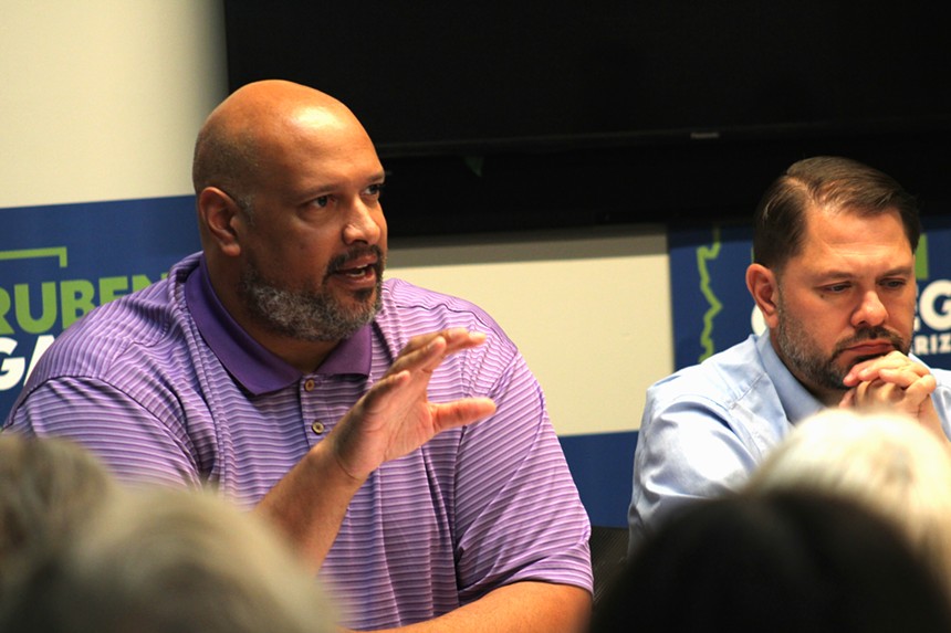 a man in a purple shirt gestures and speaks while sitting at a table