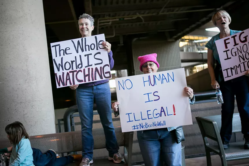 protesters hold signs that say "the world is watching" and "no human is illegal"