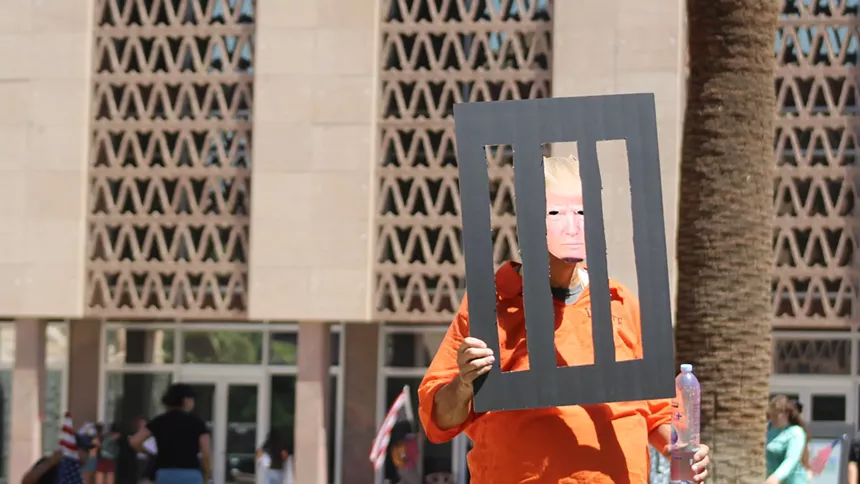 a man holds up makeshift prison bars while wearing a crude trump mask and an orange jumpsuit